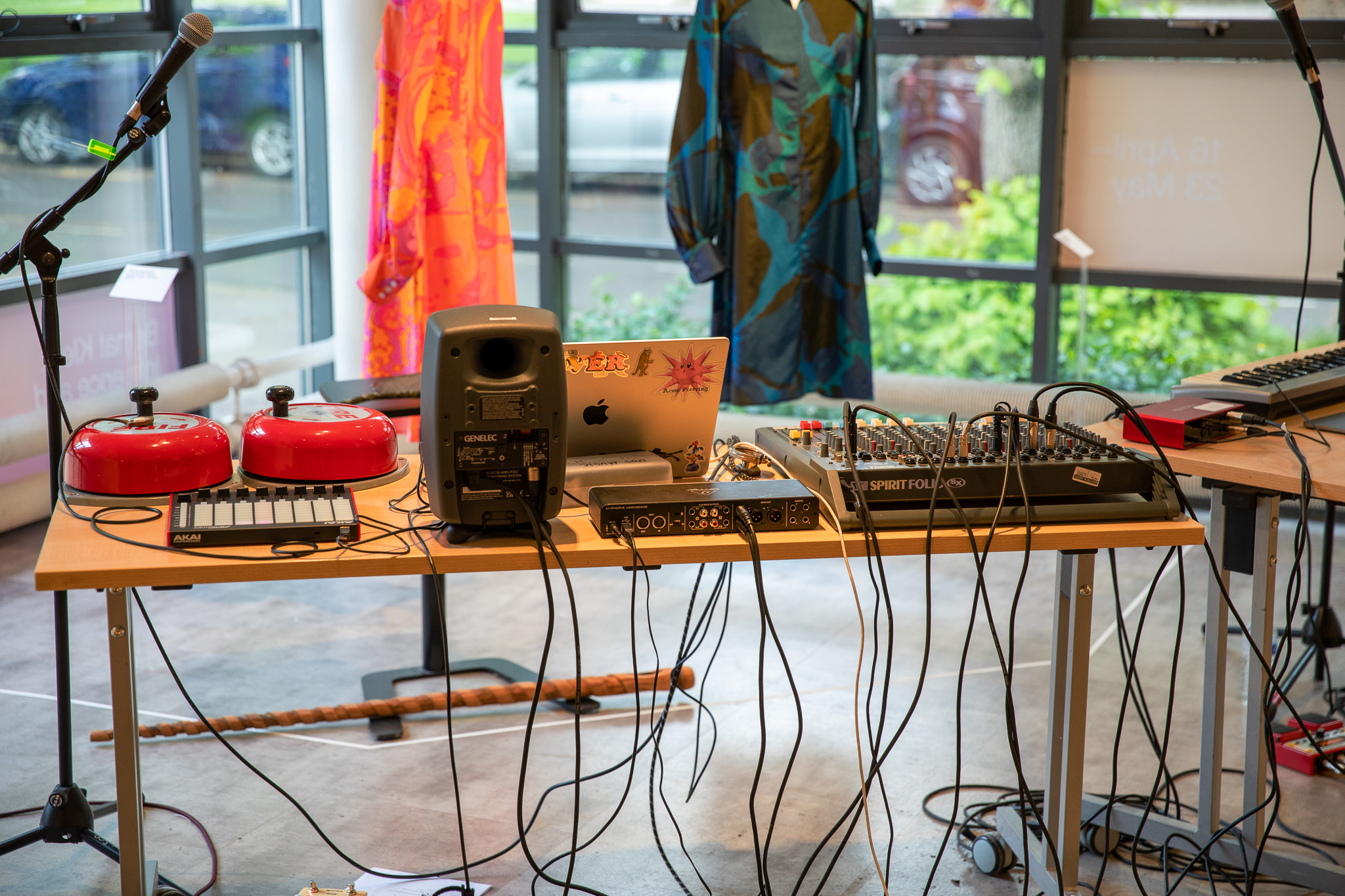 A table is set up with audio equipment including speakers, a mixer, a laptop, cables, and two red hand bells. Colourful garments hang in the background in a bright gallery space with large windows.