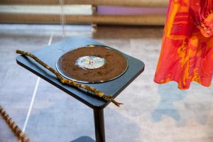 A vinyl record covered with soil sits on a small black stand beside a long stick, forming a sculptural object. Part of a brightly patterned garment hangs nearby, suggesting an exhibition setting.