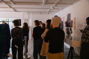 A group of people stand in a gallery facing a display, with artworks and descriptive panels mounted on the wall. A mannequin wearing a garment is positioned to the left near a roped barrier.