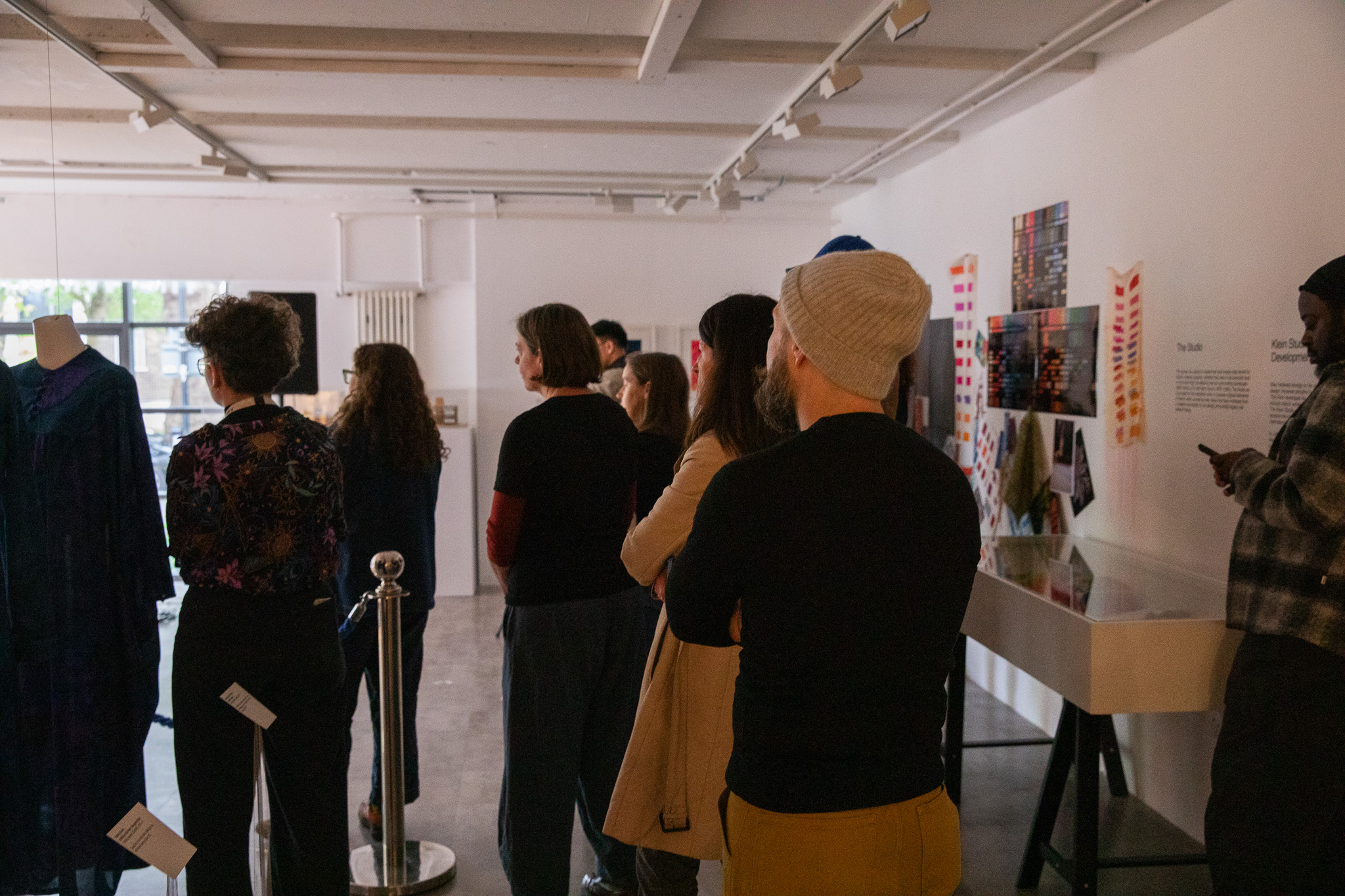 A group of people stand in a gallery facing a display, with artworks and descriptive panels mounted on the wall. A mannequin wearing a garment is positioned to the left near a roped barrier.
