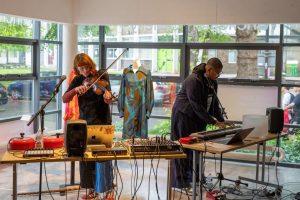 Two performers stand behind tables filled with audio equipment, with one playing a violin and the other using a keyboard and laptop. Behind them, garments are displayed on mannequins in a bright gallery space with large windows.