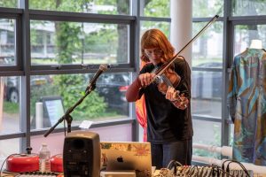 A person plays a violin beside a microphone, positioned behind a table of audio equipment including a laptop, mixer and speakers. Large windows and a mannequin displaying a patterned garment appear in the background.