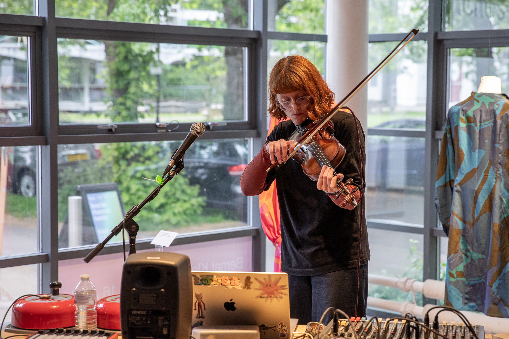 A person plays a violin beside a microphone, positioned behind a table of audio equipment including a laptop, mixer and speakers. Large windows and a mannequin displaying a patterned garment appear in the background.