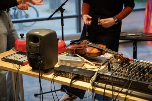 A close‑up shows a violin placed on a table among audio equipment, cables and studio devices. Two red hand bells and a speaker sit nearby, with hands interacting with cables and connectors.