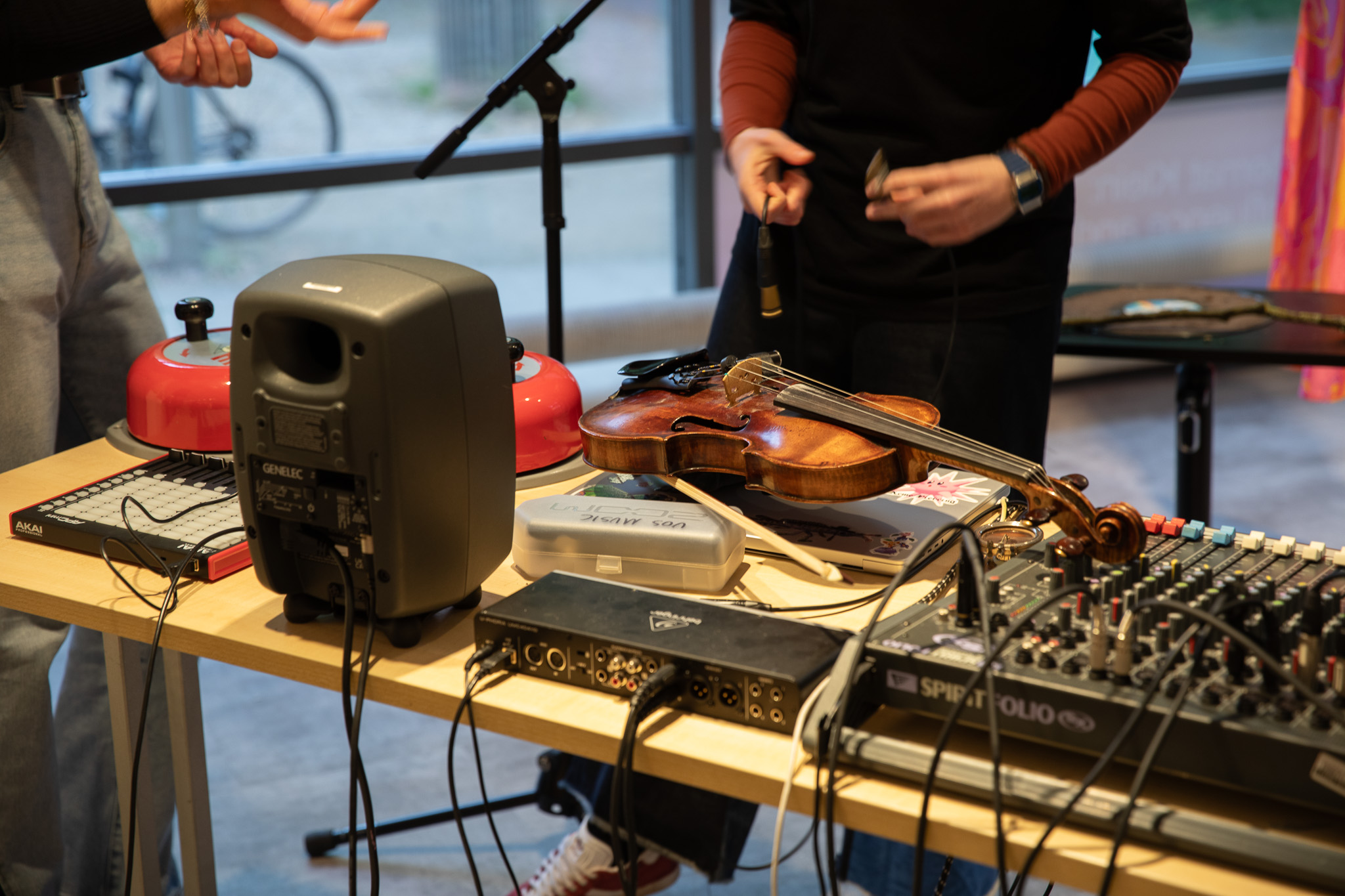 A close‑up shows a violin placed on a table among audio equipment, cables and studio devices. Two red hand bells and a speaker sit nearby, with hands interacting with cables and connectors.
