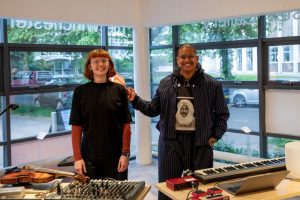 Two people stand side by side behind a table of audio equipment, including a laptop, mixer and keyboard, in a gallery space with large windows. A violin rests on the table and a mannequin displaying a patterned garment is visible in the background.