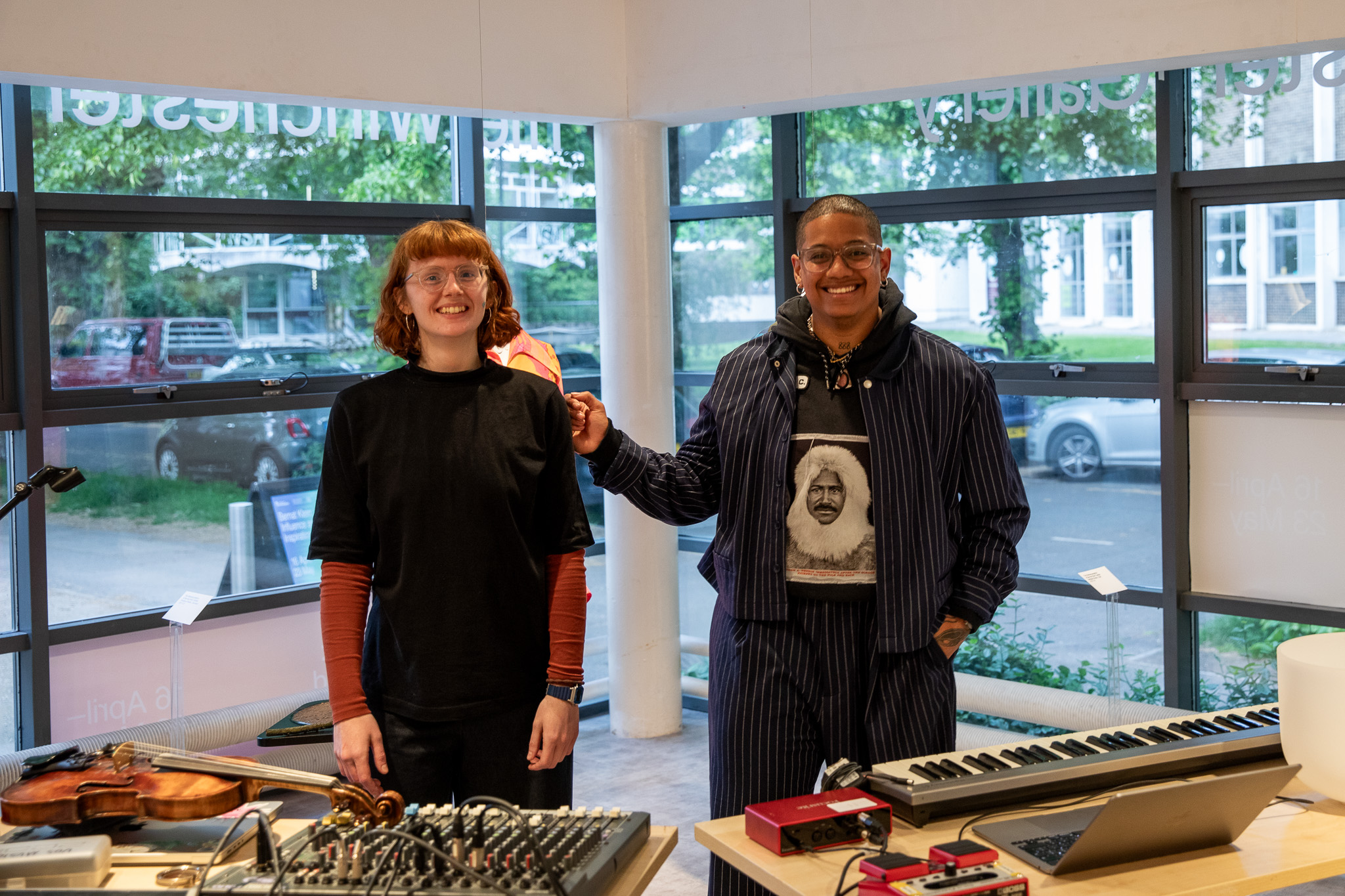 Two people stand side by side behind a table of audio equipment, including a laptop, mixer and keyboard, in a gallery space with large windows. A violin rests on the table and a mannequin displaying a patterned garment is visible in the background.