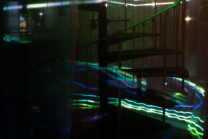 A spiral staircase is partially obscured by streaks of neon green, blue and white light projected across glass surfaces. The curved railings and steps are visible through reflections and layered visual effects.