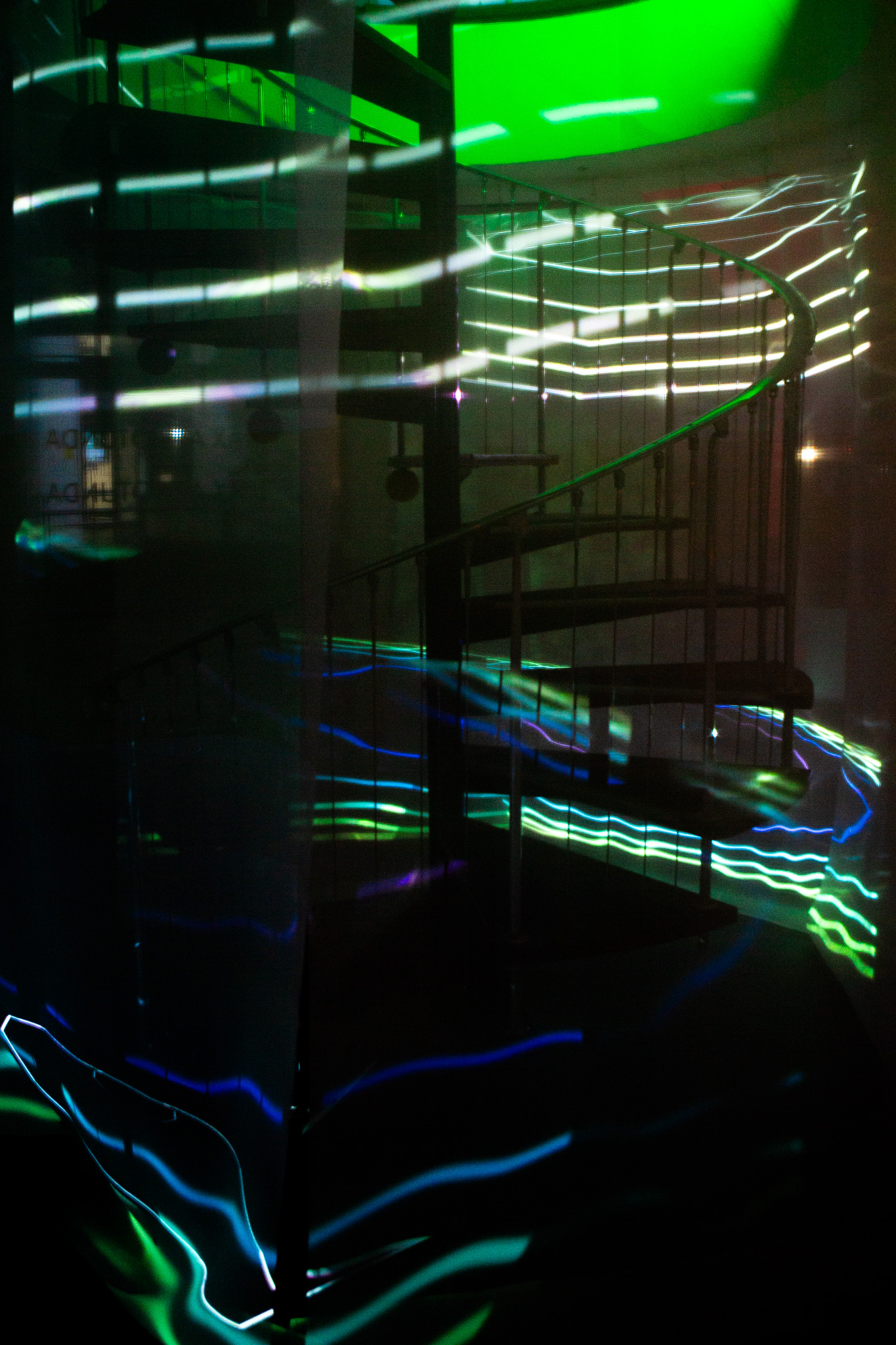 A spiral staircase is partially obscured by streaks of neon green, blue and white light projected across glass surfaces. The curved railings and steps are visible through reflections and layered visual effects.