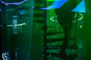 A translucent fabric or screen hangs in front of a spiral staircase, illuminated by green and blue projected shapes and lines. The layered projection creates a blurred, abstract view of the architectural structure behind it.