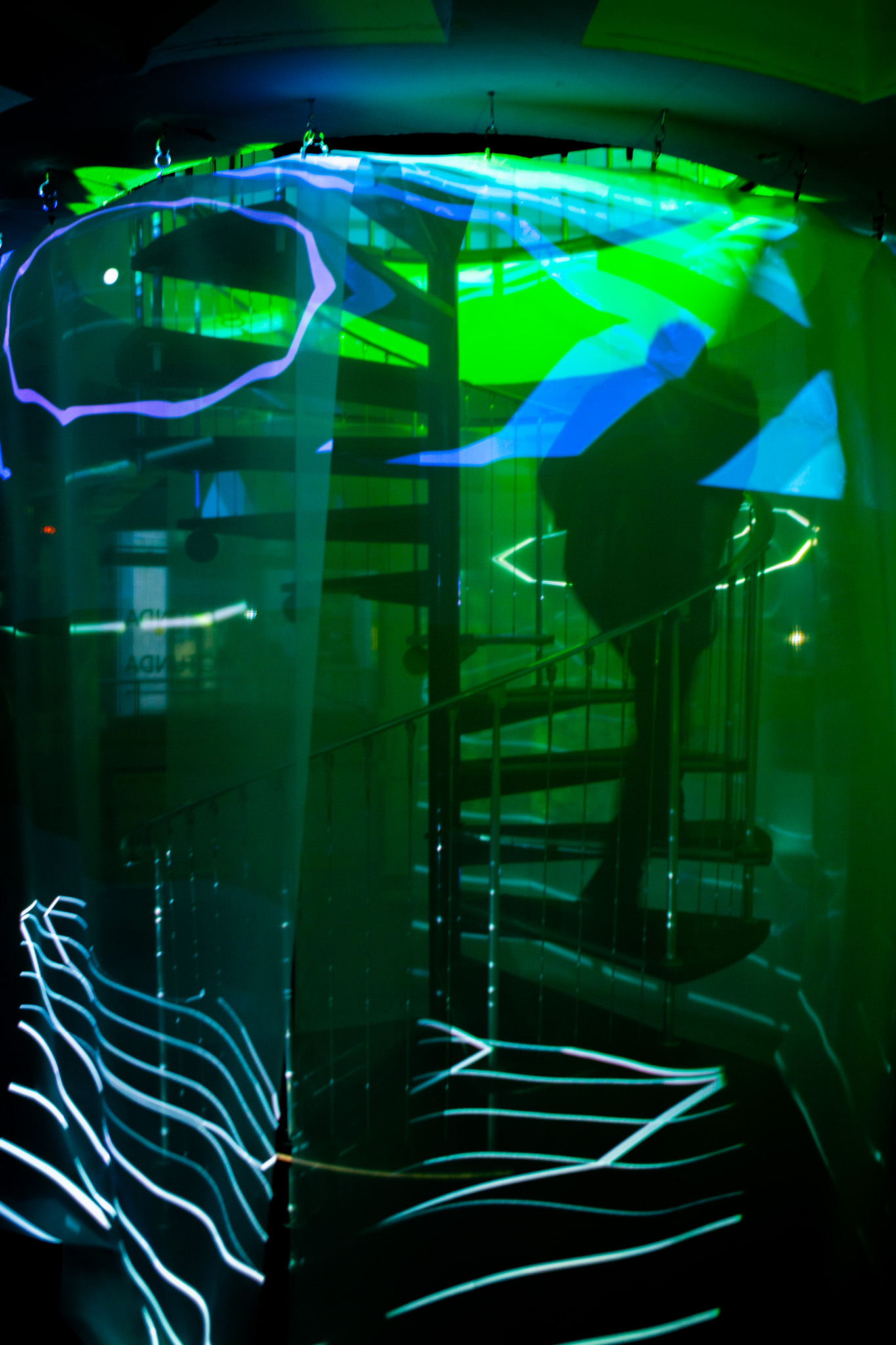 A translucent fabric or screen hangs in front of a spiral staircase, illuminated by green and blue projected shapes and lines. The layered projection creates a blurred, abstract view of the architectural structure behind it.