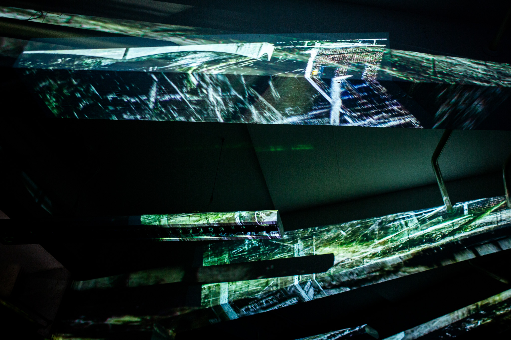 Abstract digital projections in green and white form fragmented shapes across dark surfaces, resembling distorted architectural patterns. The scene appears immersive, with overlapping layers of light and shadow.