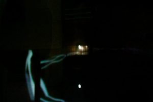 In a darkened space, faint white light traces form a circular ring on the floor while a dim rectangular glow appears in the background. The surrounding environment is mostly in shadow, with minimal visible detail.
