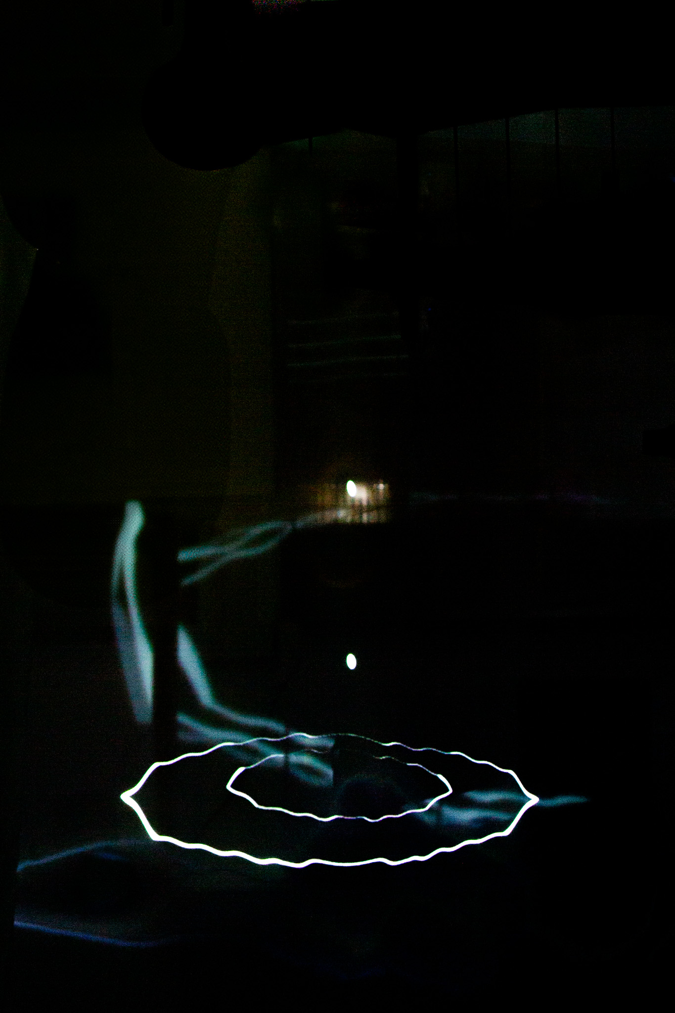 In a darkened space, faint white light traces form a circular ring on the floor while a dim rectangular glow appears in the background. The surrounding environment is mostly in shadow, with minimal visible detail.