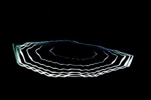 A glowing white ring composed of layered, wavy lines is projected onto a dark surface, creating a halo‑like shape. The surrounding space is almost entirely black, emphasising the luminous contours.