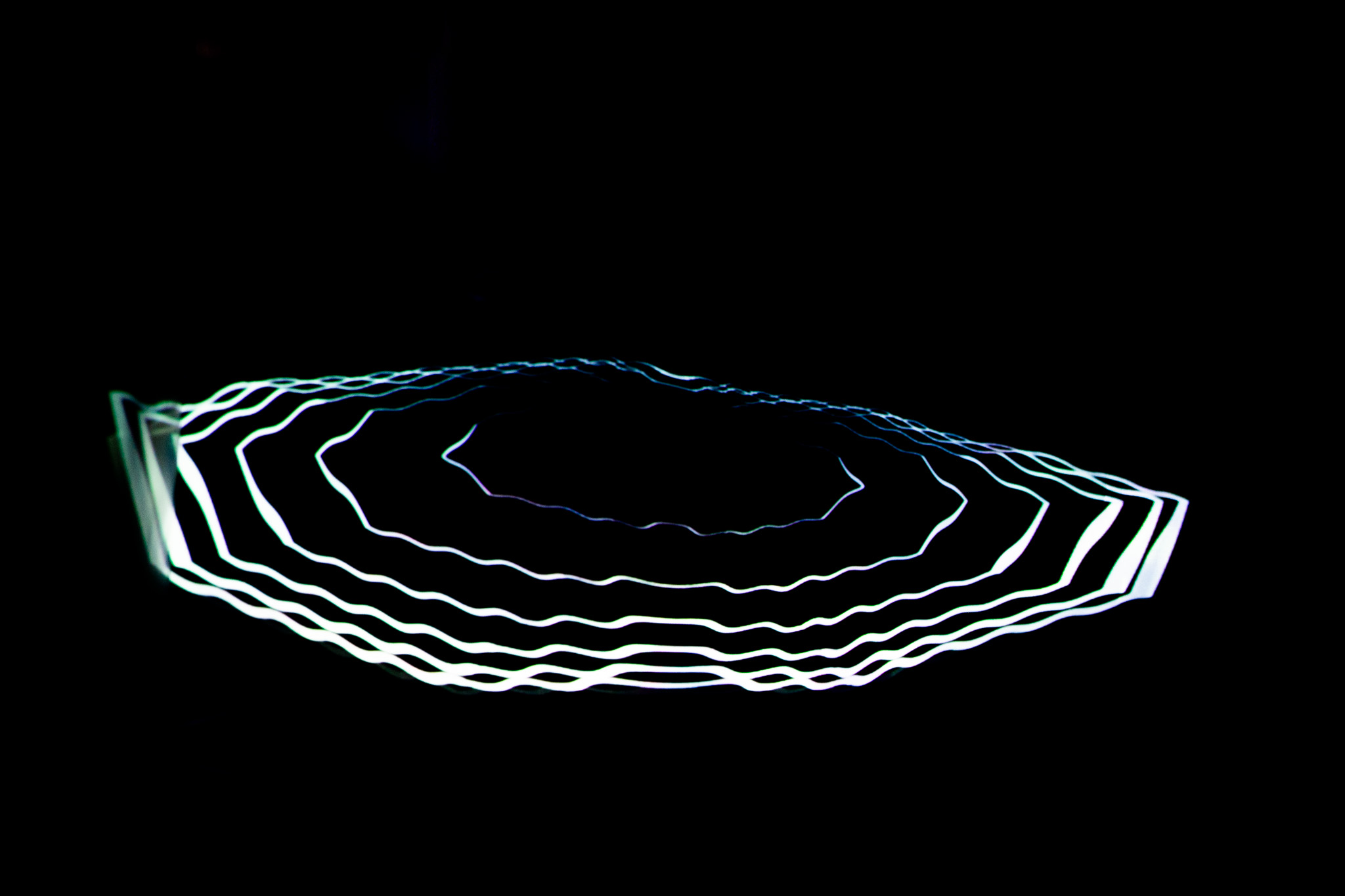 A glowing white ring composed of layered, wavy lines is projected onto a dark surface, creating a halo‑like shape. The surrounding space is almost entirely black, emphasising the luminous contours.