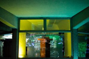 A building window is illuminated with green and yellow light, with reflections of projected lines visible on the glass. Faint text reading “RELAX AT ROTUNDA” appears across the surface.