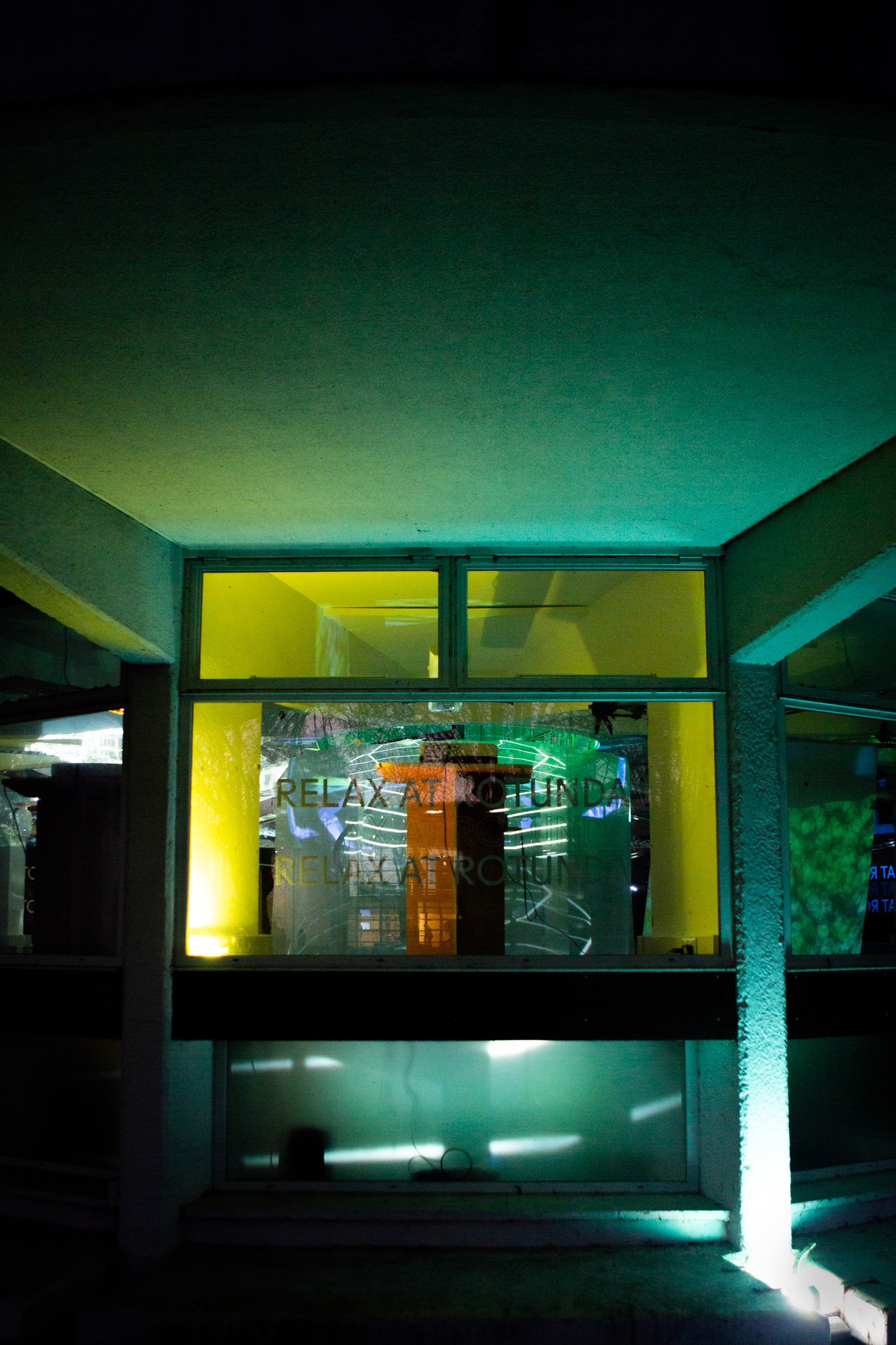 A building window is illuminated with green and yellow light, with reflections of projected lines visible on the glass. Faint text reading “RELAX AT ROTUNDA” appears across the surface.