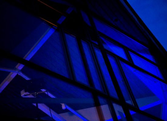 Blue light washes across a glass façade with metal framing, highlighting the geometric lines of the structure. The interior ceiling and beams are faintly visible through the tinted windows.