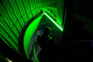 A spiral staircase is lit with vivid green light, with curved railings and steps forming a circular pattern. Thin white projected lines run along the structure while a person walks down the stairs.
