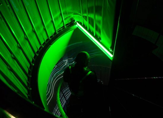 A spiral staircase is lit with vivid green light, with curved railings and steps forming a circular pattern. Thin white projected lines run along the structure while a person walks down the stairs.