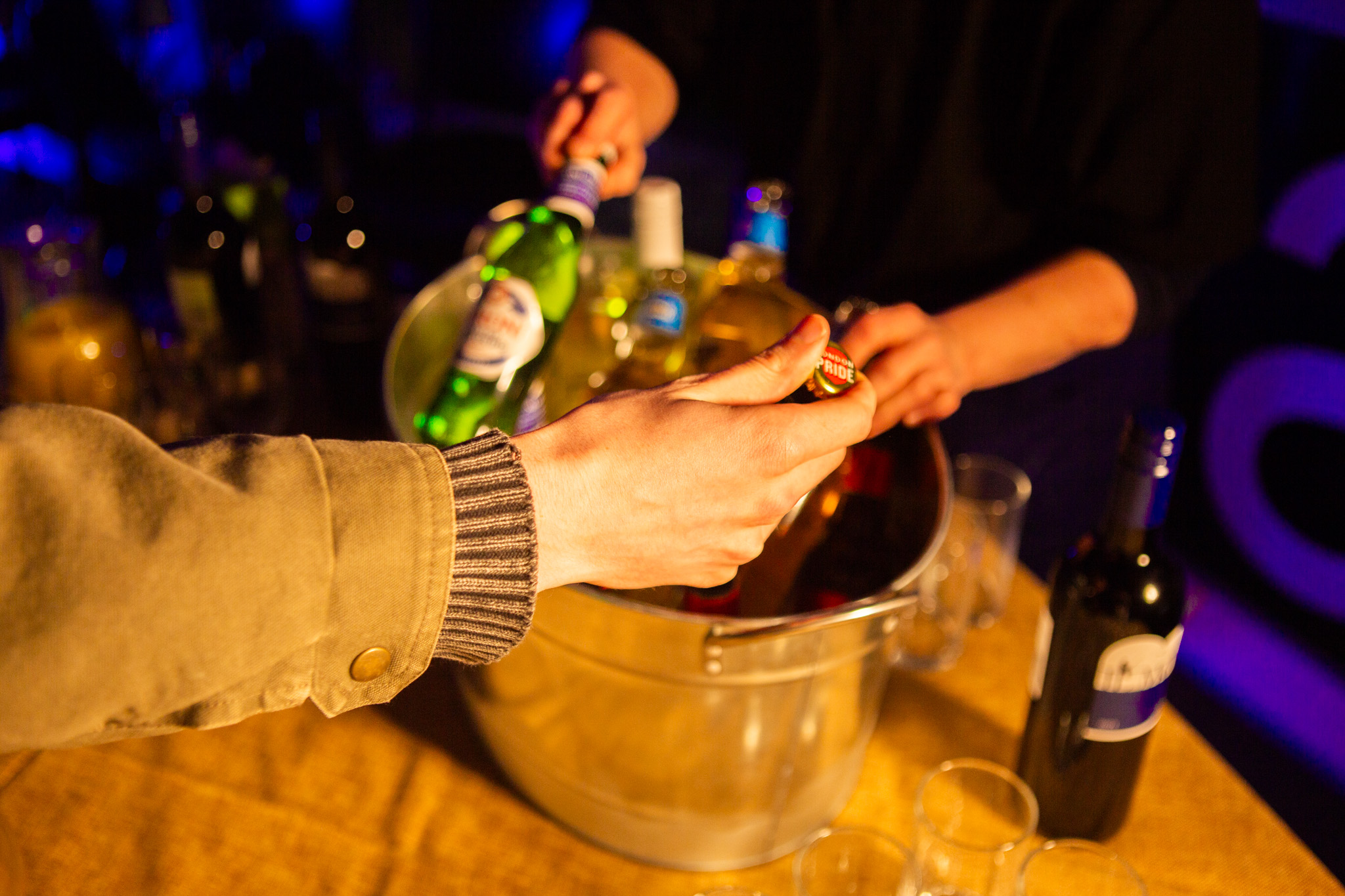 Hands open a bottle over a metal bucket filled with ice and assorted drinks on a wooden table. Warm lighting highlights glassware and bottles arranged closely together.