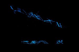 Abstract blue line projections form angular, fragmented shapes across a dark surface. The surrounding space is largely black, with the luminous lines creating a geometric pattern.