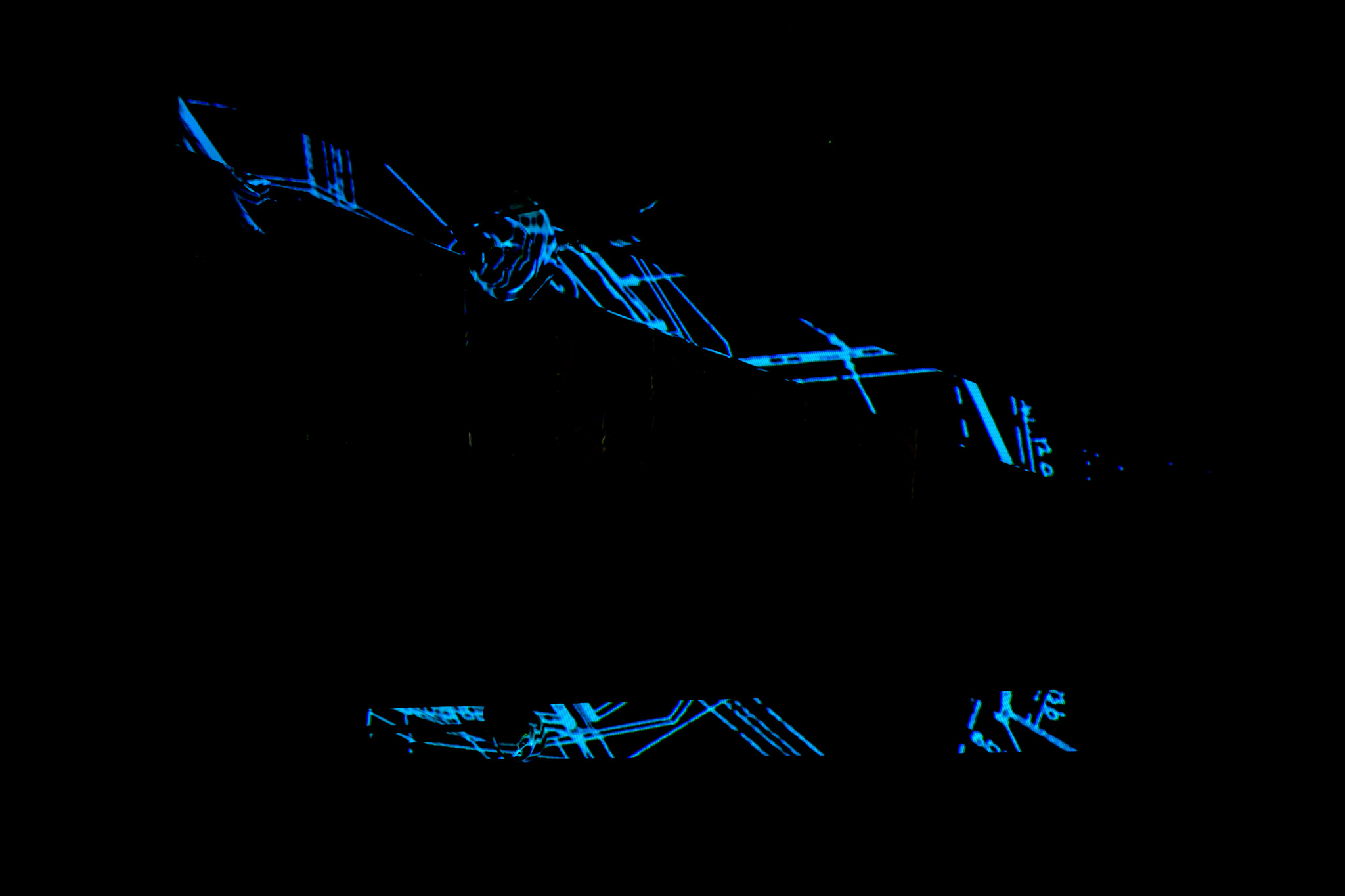 Abstract blue line projections form angular, fragmented shapes across a dark surface. The surrounding space is largely black, with the luminous lines creating a geometric pattern.