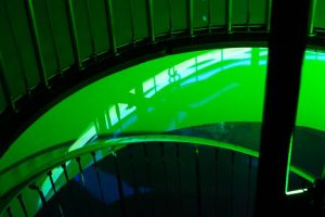 A spiral staircase is illuminated by bright green light, with curved railings and vertical bars forming a circular pattern. Reflections and shadows create layered shapes across the surfaces, and an exit sign is visible on the wall.