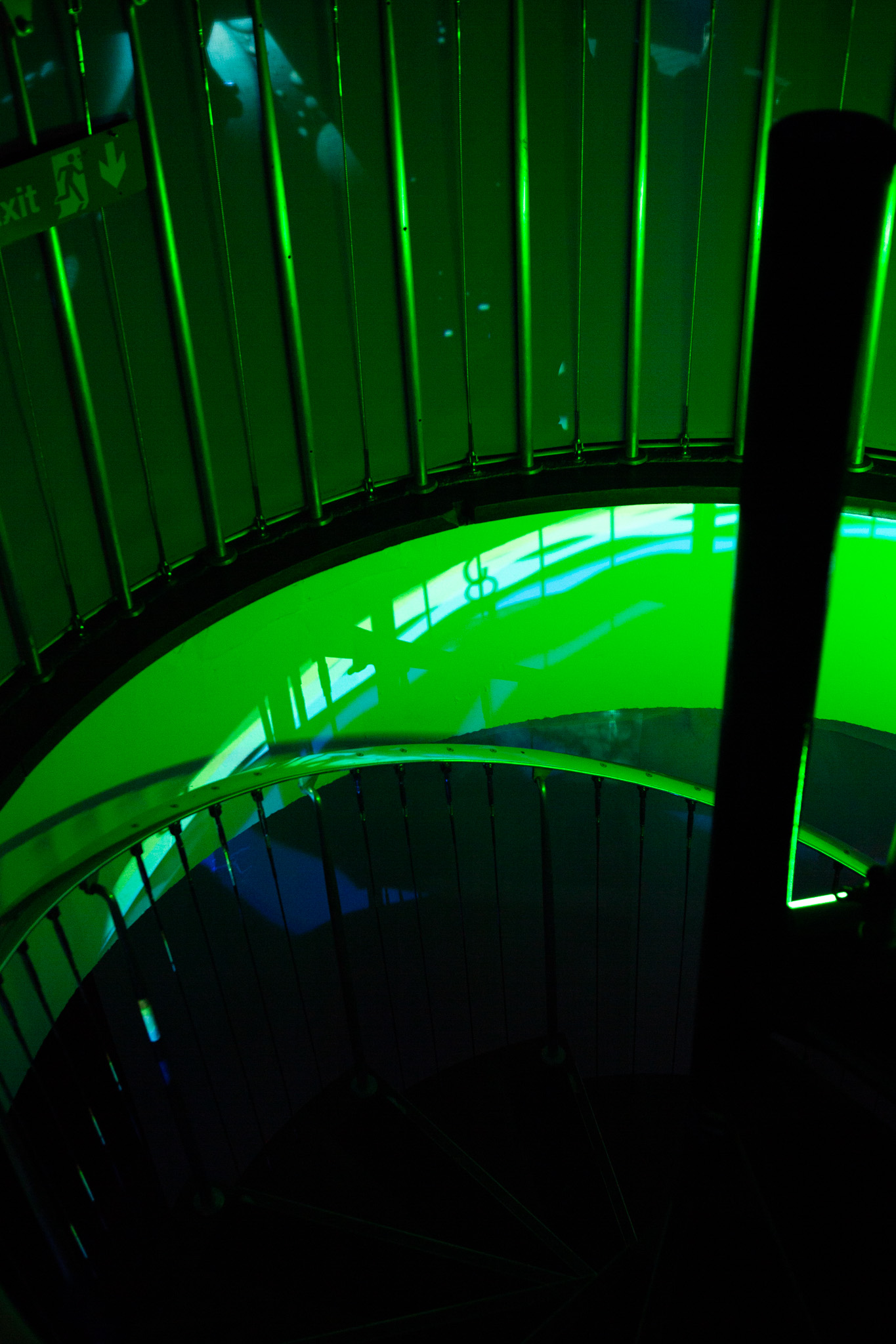 A spiral staircase is illuminated by bright green light, with curved railings and vertical bars forming a circular pattern. Reflections and shadows create layered shapes across the surfaces, and an exit sign is visible on the wall.