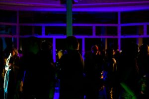 A group of people stand in a dimly lit room near large windows, with purple and blue lighting casting strong silhouettes. Some people hold drinks while others face each other in conversation.