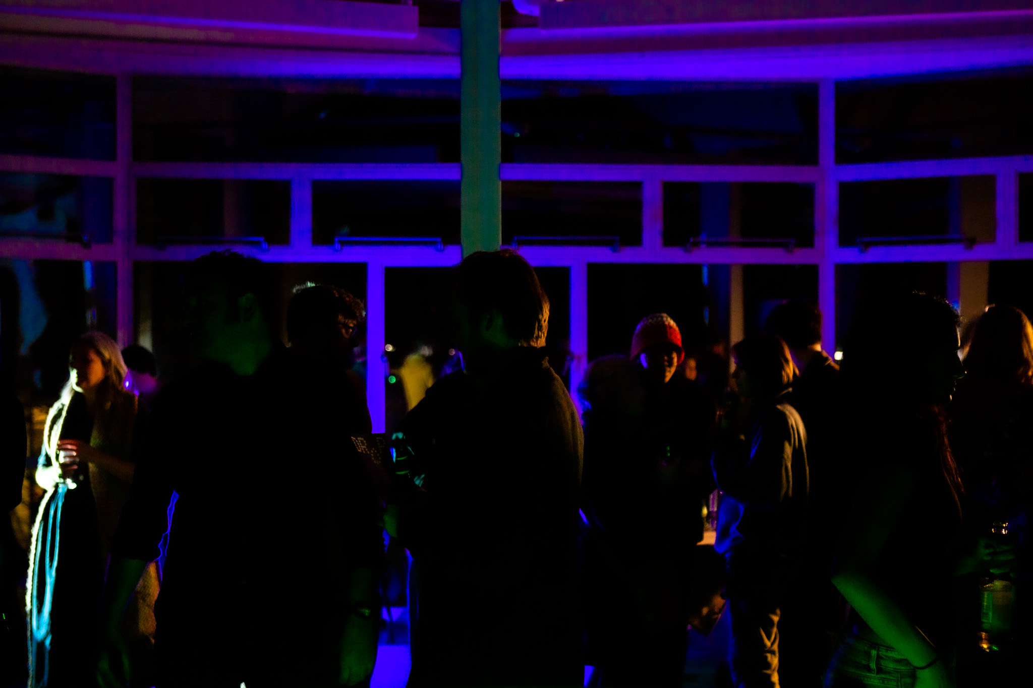 A group of people stand in a dimly lit room near large windows, with purple and blue lighting casting strong silhouettes. Some people hold drinks while others face each other in conversation.