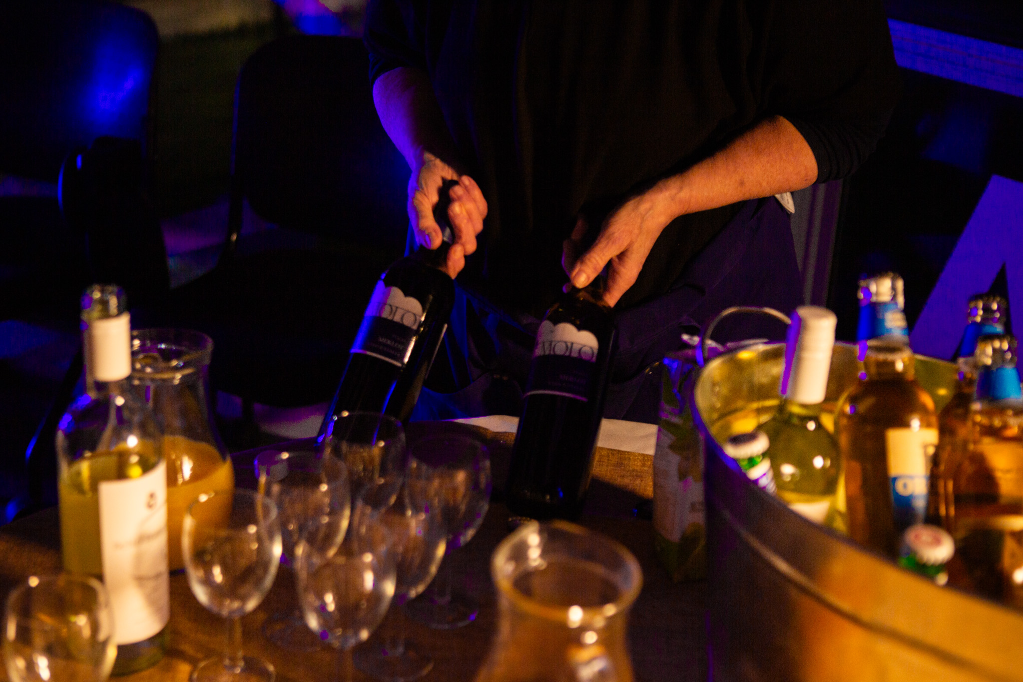 Hands hold and open bottles of wine over a table filled with glasses, a bucket of drinks and assorted bottles. Warm lighting highlights the glassware and reflective surfaces in a social setting.