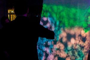 Silhouetted figures stand in a dark room illuminated by vivid purple and green projected lights, with one person reaching toward a colorful abstract display on the wall. The scene has a moody, immersive atmosphere with blurred glowing patterns and neon tones.