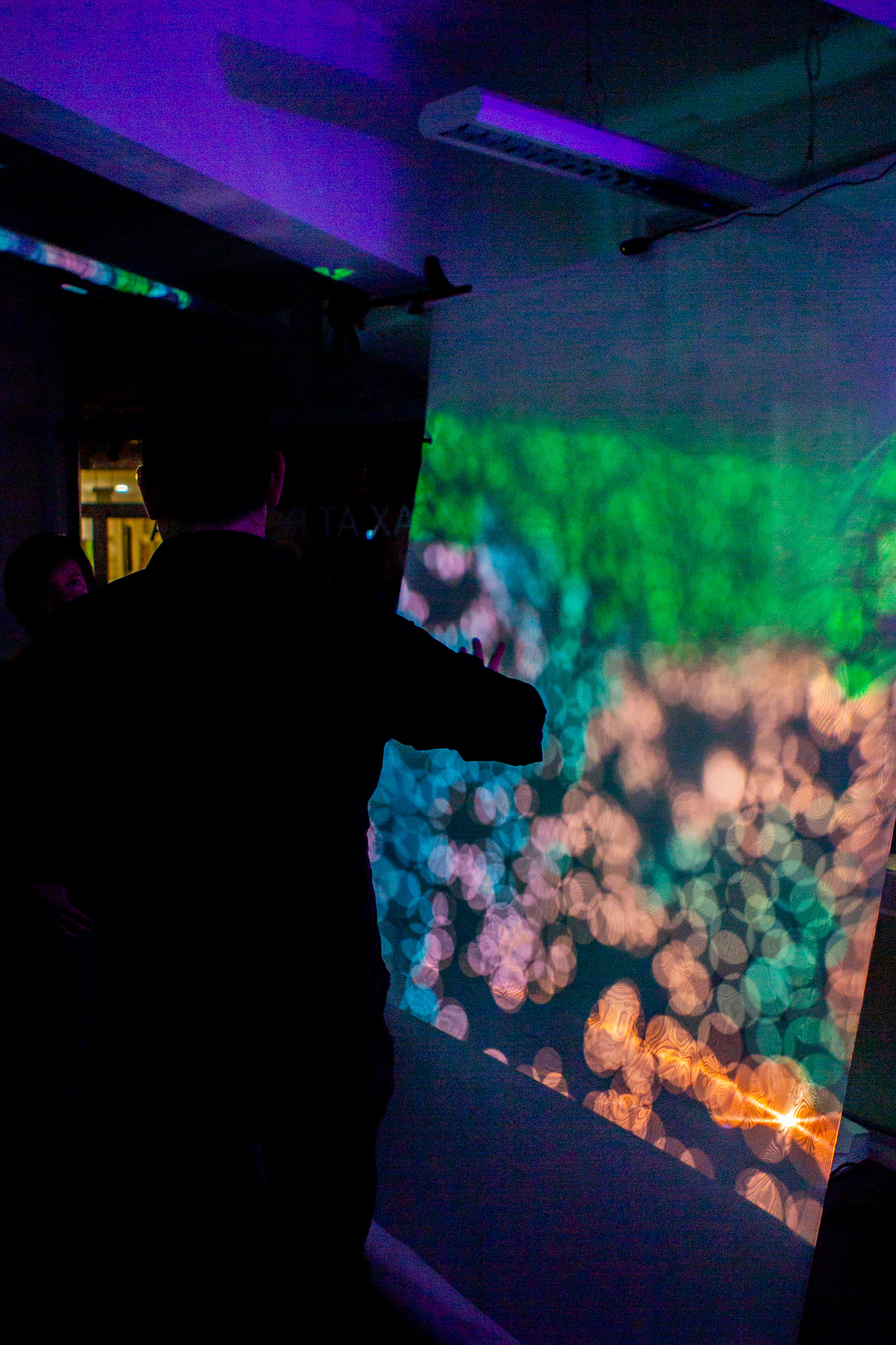 Silhouetted figures stand in a dark room illuminated by vivid purple and green projected lights, with one person reaching toward a colorful abstract display on the wall. The scene has a moody, immersive atmosphere with blurred glowing patterns and neon tones.