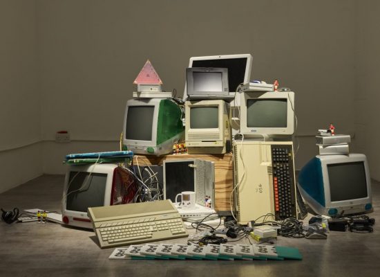 A gallery display shows a stack of vintage computers and monitors arranged in a sculptural installation, with keyboards, cables and small objects placed around the base. The mix of older desktop machines and accessories creates a layered composition against a plain white wall.