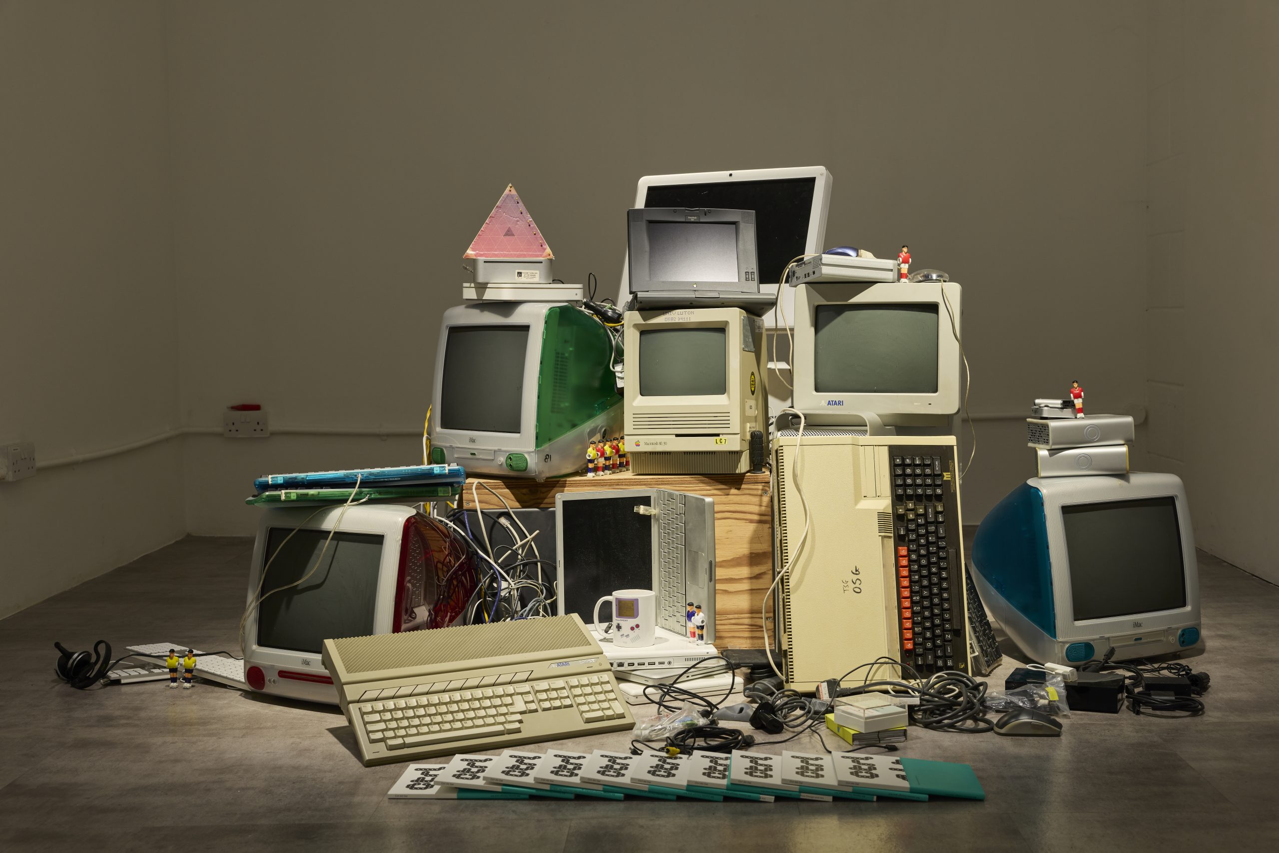 A gallery display shows a stack of vintage computers and monitors arranged in a sculptural installation, with keyboards, cables and small objects placed around the base. The mix of older desktop machines and accessories creates a layered composition against a plain white wall.