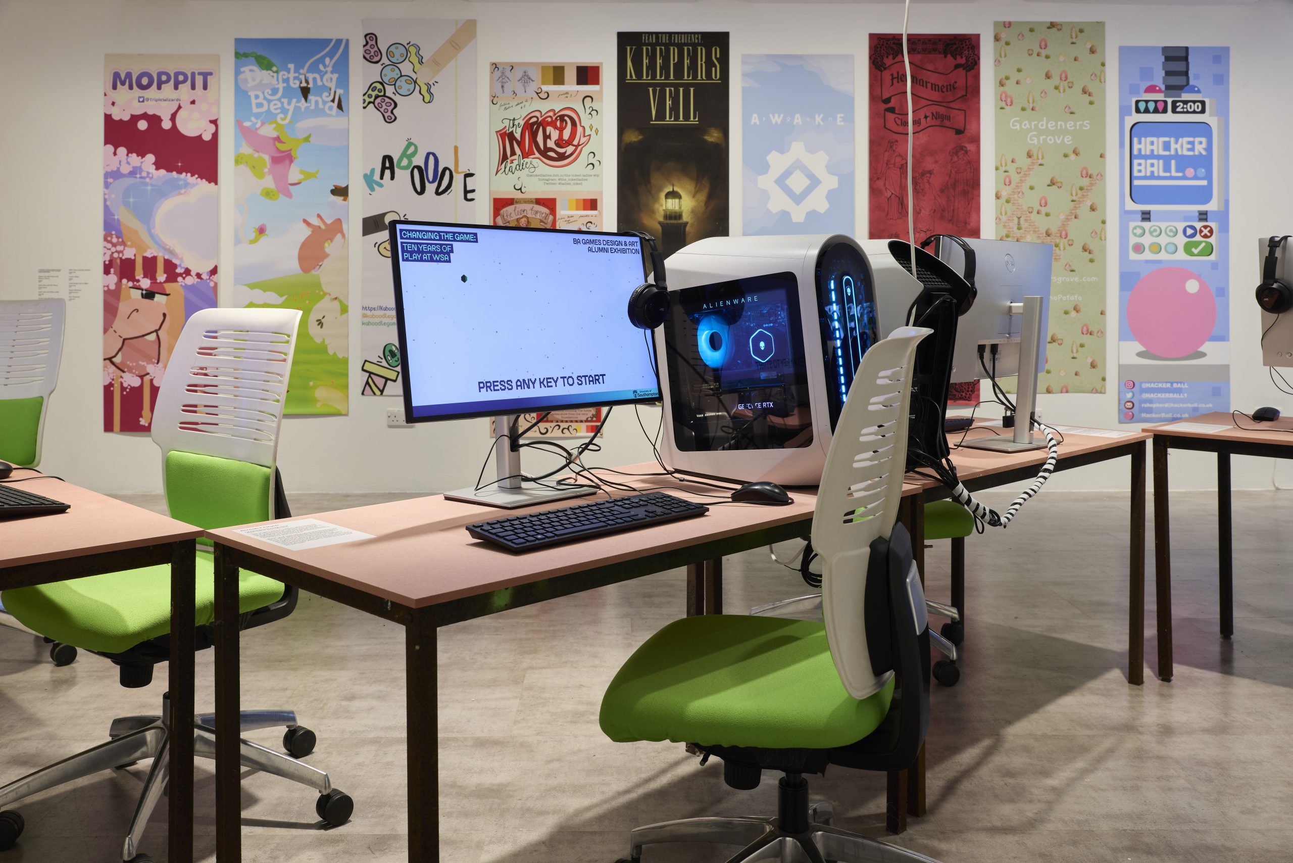 A workspace displays several computers and monitors on long tables with green chairs, including a tower with blue internal lighting and a screen reading “Press any key to start.” Colourful illustrated posters line the wall behind, forming a vibrant backdrop.