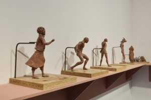 A row of clay sculptures depicts human and animal‑like figures mounted on wooden bases, some connected to metal rods extending from their backs. The figures are arranged along a shelf against a plain wall, progressing from clothed to more abstract forms.