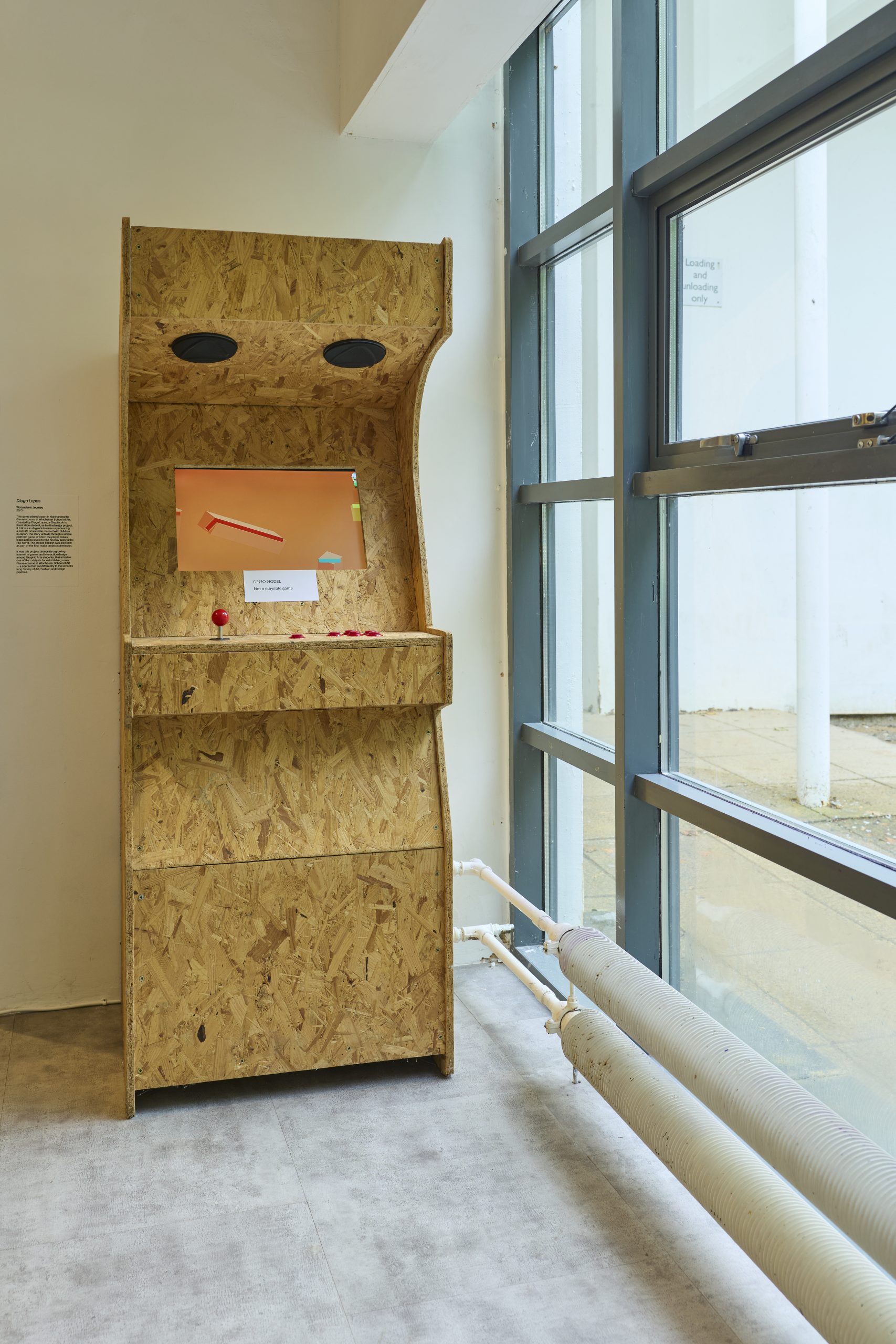 A wooden arcade‑style cabinet with a small screen and joystick stands beside a tall window, showing a simple game on an orange background. Pipes run along the wall and floor, and natural light enters from the window.