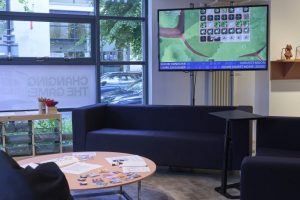 A lounge area with sofas and a low table is arranged around a large screen displaying a pixel‑style farming game. Printed materials and small cards are spread on the table, and large windows reveal trees and parked cars outside.