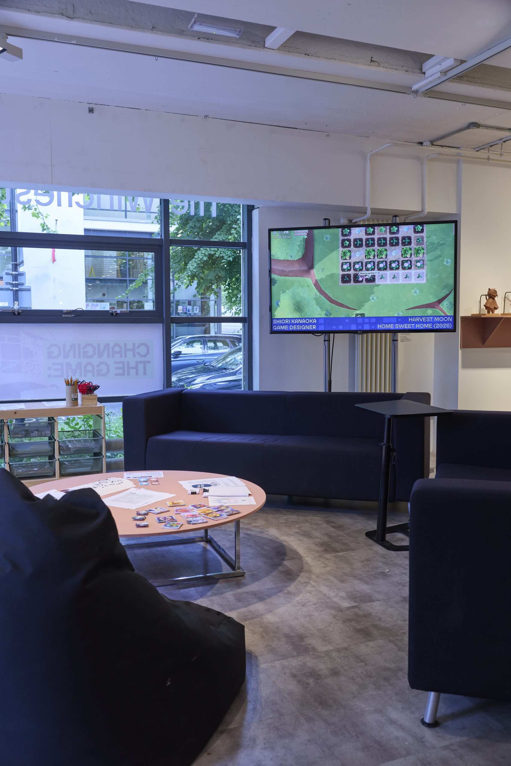 A lounge area with sofas and a low table is arranged around a large screen displaying a pixel‑style farming game. Printed materials and small cards are spread on the table, and large windows reveal trees and parked cars outside.