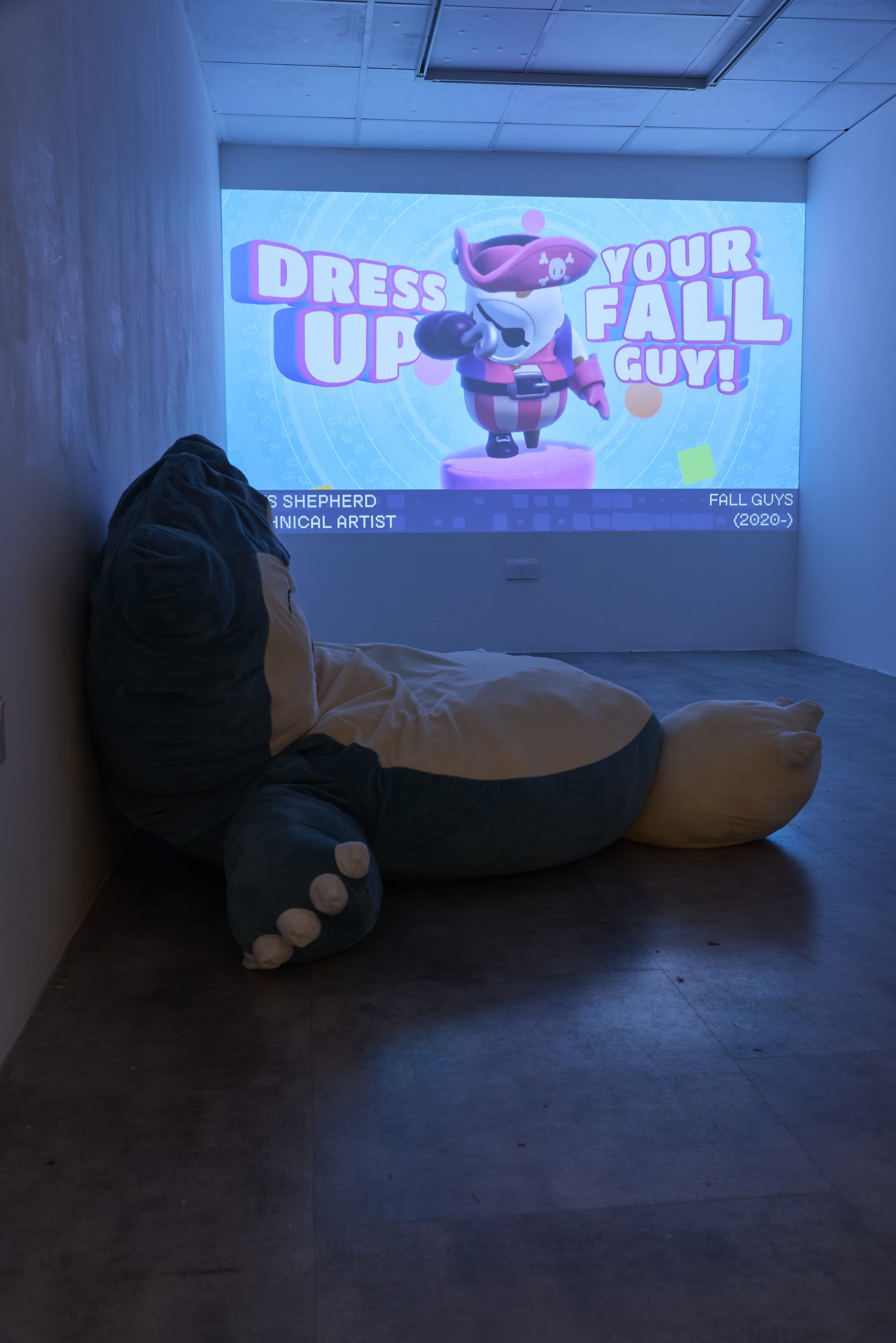 A large projection shows a colourful screen reading “DRESS UP YOUR FALL GUY!” with a cartoon character in a pirate outfit in a dark room. In the foreground, a large plush seat rests on the floor facing the projection, suggesting an interactive viewing area.