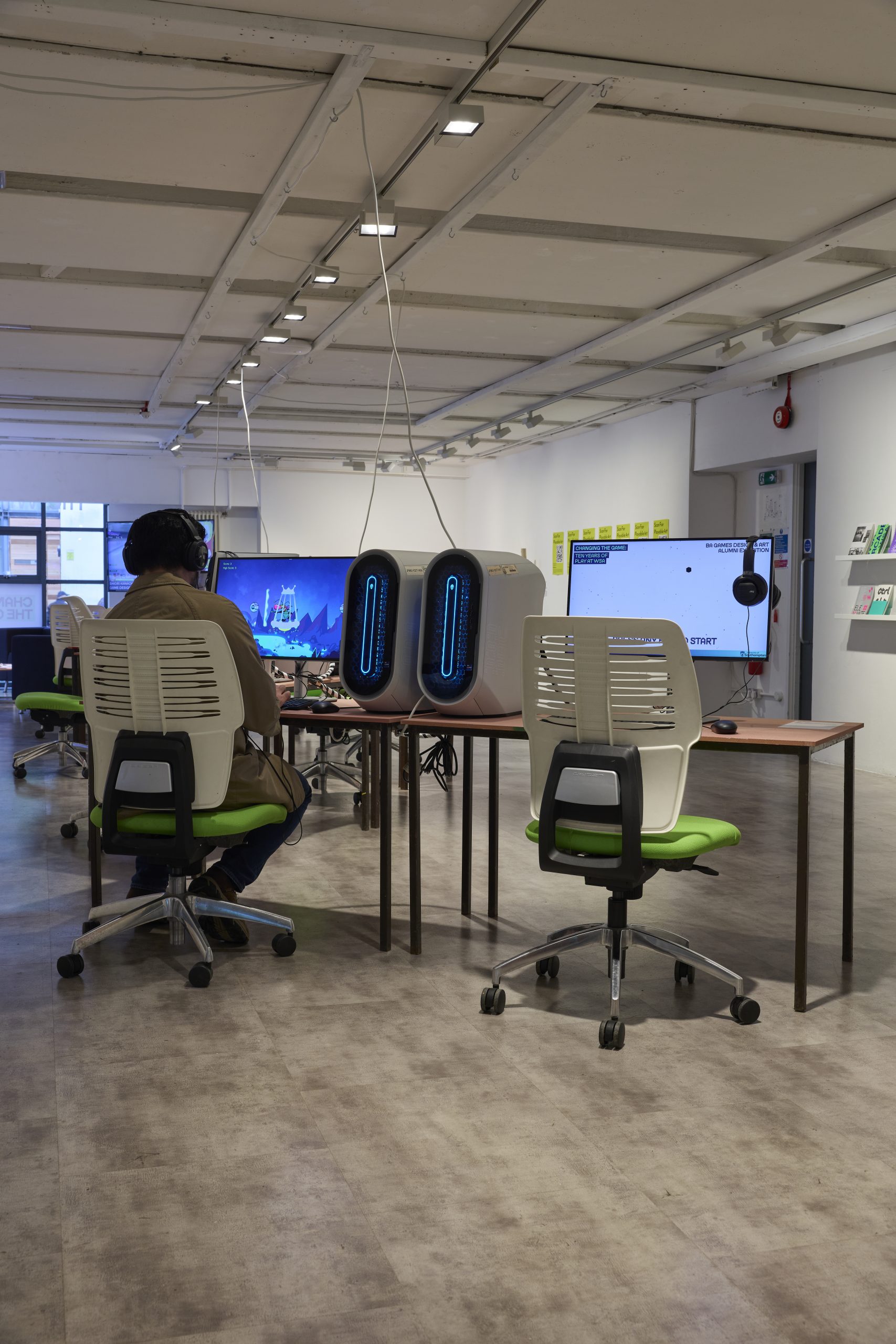 A person sits at a long table using a computer with headphones in a bright gallery workspace, surrounded by monitors and gaming PCs with blue lighting. White chairs and neat rows of desks extend through the space beneath exposed ceiling beams and track lighting.