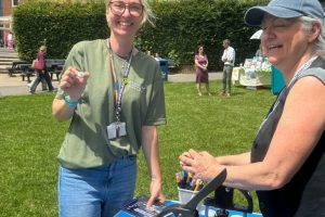 Two people take part in a button-making activity at an outdoor community event. A table with craft materials and equipment is set up on a lawn with buildings and visitors in the background.