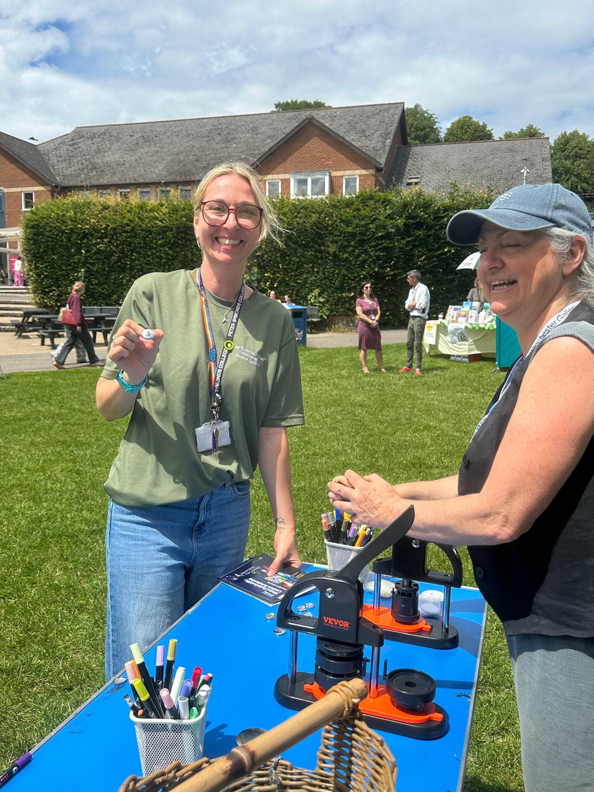 Two people take part in a button-making activity at an outdoor community event. A table with craft materials and equipment is set up on a lawn with buildings and visitors in the background.