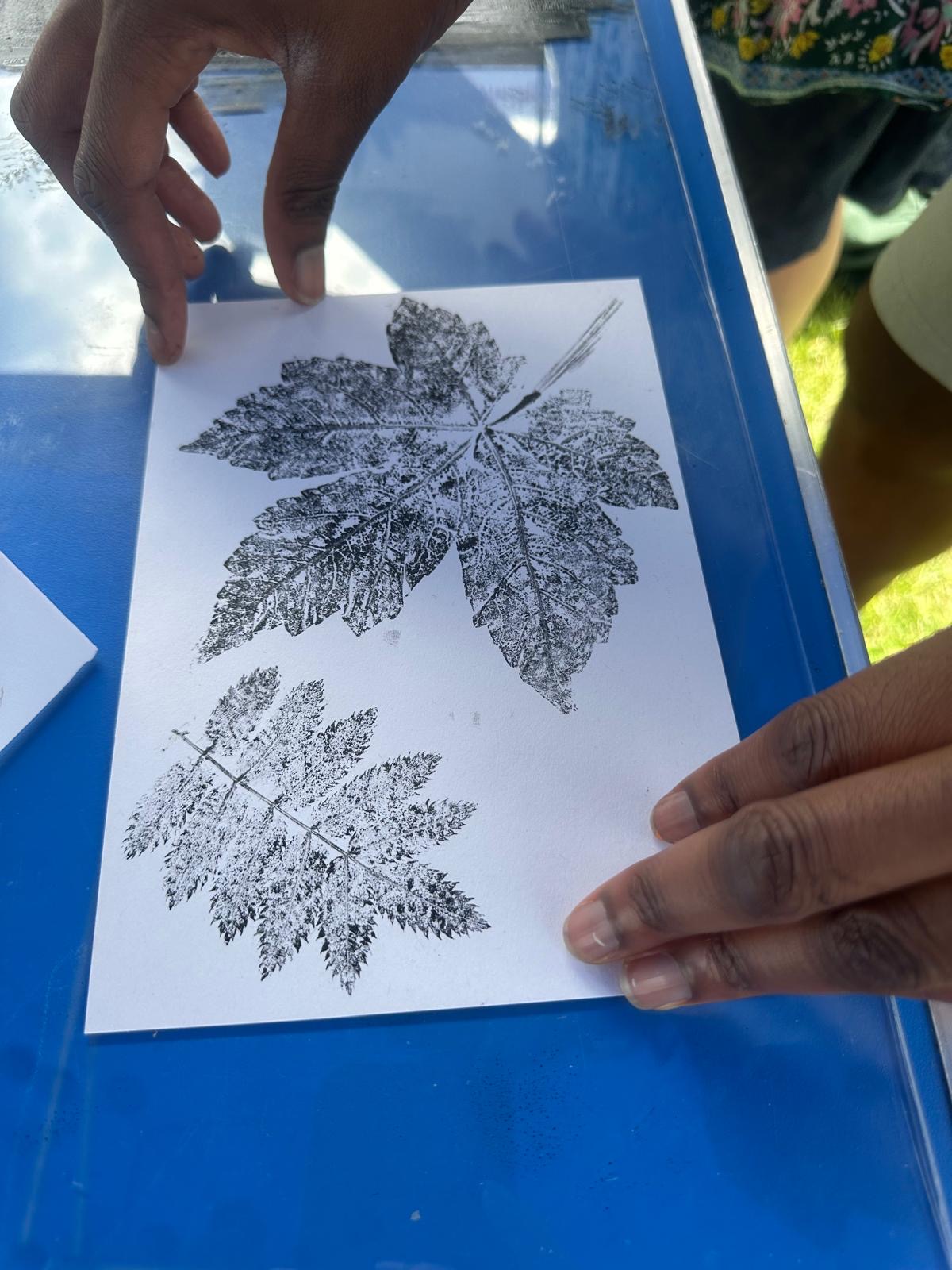 Hands hold a sheet of paper showing detailed leaf prints created using a nature-printing technique. The black ink impressions highlight the veins and shapes of two different leaves.