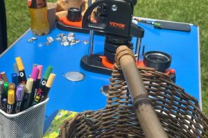 A tabletop display at an outdoor event features a button-making press, coloured pens and a woven basket. The activity materials are arranged on a blue table set on a grassy lawn.