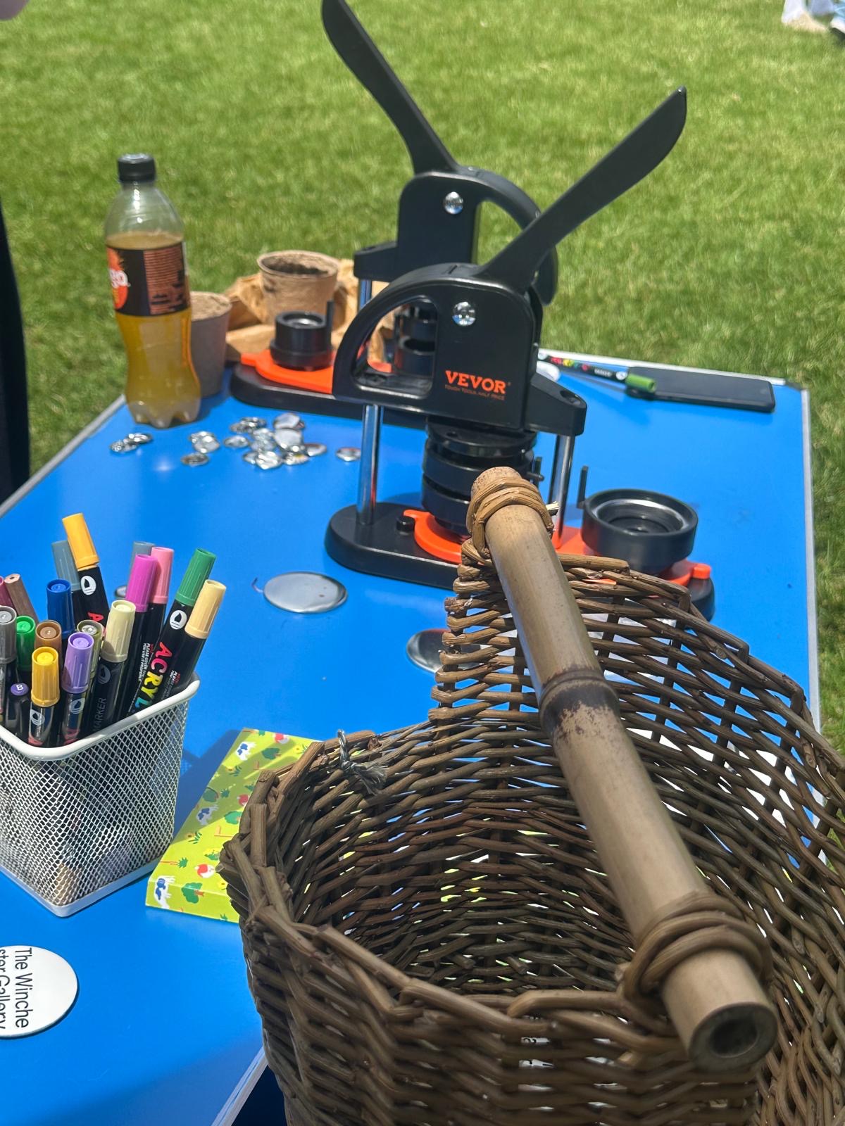 A tabletop display at an outdoor event features a button-making press, coloured pens and a woven basket. The activity materials are arranged on a blue table set on a grassy lawn.