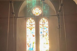 Bell-ringing ropes hang in front of a stained-glass church window illuminated by bright sunlight. Green reflections from the stained glass are cast onto the stone arch and wall above.